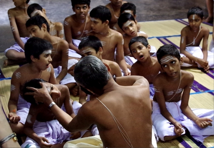 写真１　インド南部ケーララ州Thrissurにある伝統的なヴェーダの学校Brahmaswam Madhomにて。唱えるマントラの三種のアクセントを、師が弟子の頭を上げ下げしたり曲げたりしながら体で覚え込ませている。一人反対を向いている子供もいるが…。